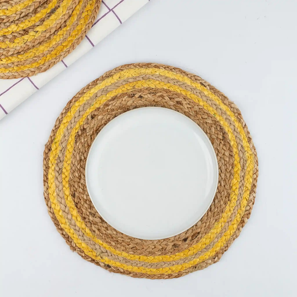 A round woven placemat with concentric rings of natural jute, tan, and mustard yellow, shown on a white surface with a white plate placed in the center.