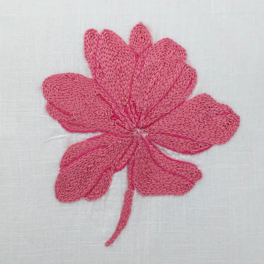 A pink knitted flower applique with six petals and a long stem, displayed on a textured white fabric background.