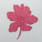 A pink knitted flower applique with six petals and a long stem, displayed on a textured white fabric background.