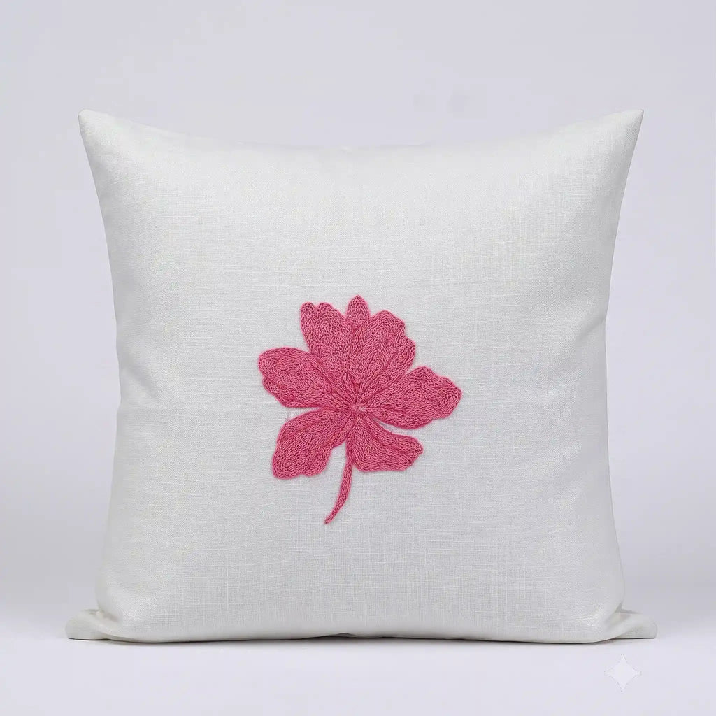 A square off-white linen throw pillow featuring a prominent pink embroidered hibiscus flower in the center, set against a clean light gray background.
