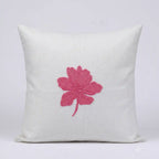 A square off-white linen throw pillow featuring a prominent pink embroidered hibiscus flower in the center, set against a clean light gray background.