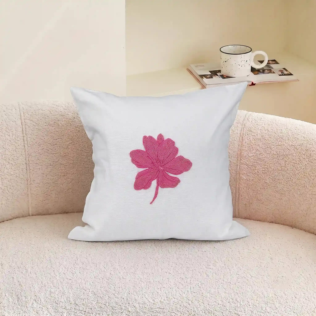 A square white throw pillow with a pink embroidered leaf design, resting on a textured beige sofa with a mug and magazine in the background.