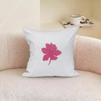 A square white throw pillow with a pink embroidered leaf design, resting on a textured beige sofa with a mug and magazine in the background.